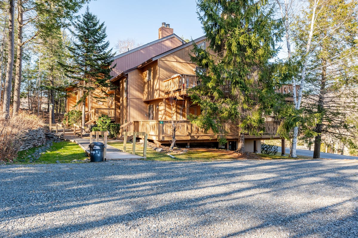 Wood-sided condo building among spruce and birch trees, with a gravel parking area in front